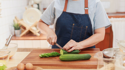 Portrait of pensioner mature woman chopping cucumber for vegetable salad dinner. Senior retired influencer woman making healthy food in the kitchen at home. Elder people lifestyle concept