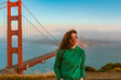 A young woman in a green hoodie stands on a hill overlooking the Golden Gate Bridge during sunset, San Francisco