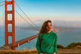 Fototapeta  - A young woman in a green hoodie stands on a hill overlooking the Golden Gate Bridge during sunset, San Francisco