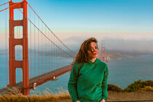 A Young Woman In A Green Hoodie Stands On A Hill Overlooking The Golden Gate Bridge During Sunset, San Francisco