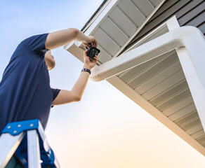 Technician on ladder installing and adjusting wireless security video camera underneath a roof for private house, monitor and recorder. Set up outdoor surveillance system. Safety and privacy concept.