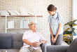 © anusak - senior man at the nursing home concept. Nurse is with an elderly man sitting on armchair in living room at the care center nursing home looking together