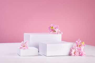 Three different square white podiums for display cosmetic produce or goods with fresh spring flowers and soft light pastel pink background.