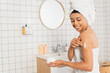 smiling young african american woman applying cream on arm in bathroom