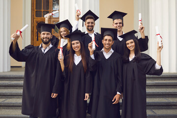 Wall Mural - Group portrait of happy graduates with honors near university. Smiling students in traditional black gowns and academic caps holding diplomas standing and looking at camera after graduation ceremony