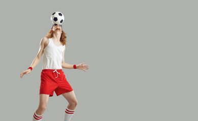 Wall Mural - Funny skinny man balancing a football on his forehead isolated on a grey copy space background. Slim sports fan in a retro white tank top and red shorts doing a nose and head stall with a soccer ball