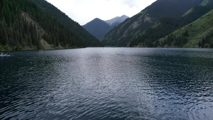 Wall Mural - Kolsai mountain lake and green forest. Top view. People swim in boats and catamarans. They rest on the green meadows. The sun's rays shine on water. Clear water and tall coniferous trees. Kazakhstan