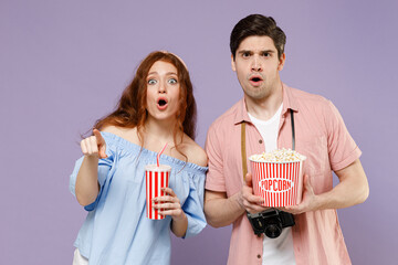 Two confused traveler tourist woman man couple in shirt hold takeaway popcorn bucket soda point finger camera isolated on purple background Passenger travel abroad weekends getaway Air flight concept.