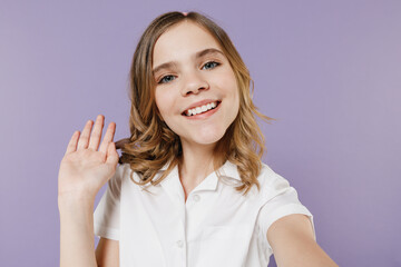 Close up fun little kid girl 12-13 years old in white shirt doing selfie shot on mobile phone waving hand say hello isolated on purple background children studio portrait. Childhood lifestyle concept.