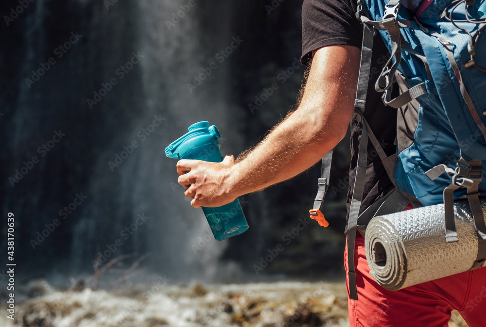 Hand close up with drinking water bottle. Man with backpack dressed in active trekking clothes touristic staying near mountain river waterfall and enjoying Nature. Traveling, trekking concept image - obrazy, fototapety, plakaty 