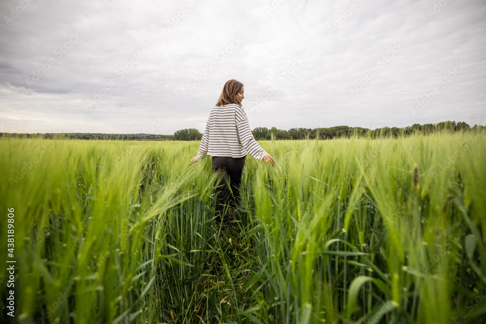 Happy woman walking in green rye field and enjoys calm nature. Freedom and meditation concept.  - obrazy, fototapety, plakaty 