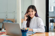 © amnaj - Asian businesswoman sitting at the office and talking on the phone working on document with a laptop at the desk. Looking at the camera.