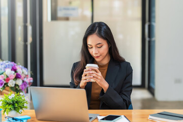 Wall Mural - Front view of a beautiful businesswoman drink coffee sitting at the office with a laptop on the table.