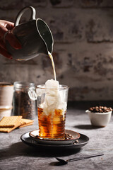 Female hand pouring milk into a glass with ice cubes and coffee