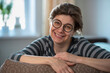 © Zadvornov - Smiling mature middle-aged woman sitting at home on the couch. Happy lady 50 years old, with glasses.