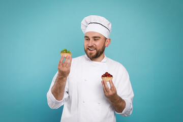 Wall Mural - Happy professional confectioner in uniform holding delicious tartlets on light blue background