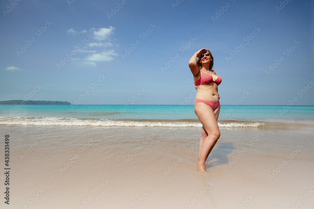 Full body of smiling overweight woman covering face from sunlight while standing on sandy beach near waving sea - obrazy, fototapety, plakaty 