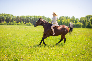 A young rider woman blonde with long hair in a dress riding gallop on brown horse on a field and forest background, Russia