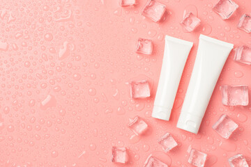 Top view photo of two different sizes white cream tubes without label ice cubes on the right and drops on isolated pastel pink background with copyspace on the left