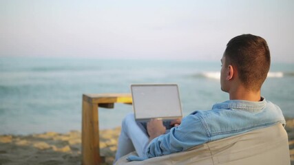 Young successful man freelancer typing and coding on laptop while lying on a lazy bag on the tropical beach at sunrise, working remotely during summer vacation. Distance and pandemic work concept