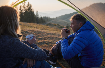 Wall Mural - Close up view from inside tent of couple of friends sitting inside tent and drinking hot tea while looking at beautiful environment. Camping in the mountains at sunny day.