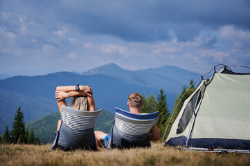 Wall Mural - Rear view of tourists pair basking in the sun, sitting in trekking chairs near tent in their campsite and admiring surrounding mountain landscape. Concept of hiking, camping and relationships.