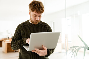 Young ginger man working with laptop while standing in office