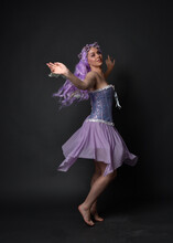 Full Length Portrait Of A Purple Haired  Girl Wearing Fantasy Corset Dress With Fairy Wings And Flower Crown.  Standing Pose Against A Dark Studio Background.