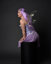 Full Length Portrait Of A Purple Haired  Girl Wearing Fantasy Corset Dress With Fairy Wings And Flower Crown.  Seated Pose Against A Dark Studio Background.