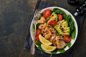 Wall Mural - Fresh green salad with grilled chicken fillet, spinach, tomatoes, avocado, lemon and black sesame seeds in white bowl on old wooden dark table background. Nutrition Diet Concept. Top view .