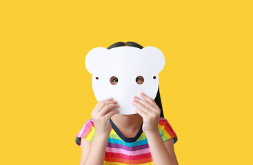 Wall Mural - Little child girl holding blank white animal paper mask fronting her face isolated on yellow background. Idea and concept for kid dressed up playing animal face.