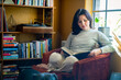 © Inti St. Clair - Woman sitting in orange chair by window and bookshelf reading a book