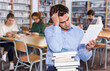 © JackF - Portrait of tired upset adult man sitting with pile of books in public library, browsing one of them
