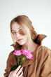 Thoughtful young woman in jacket with brown birthmark on face and pink flowers in hand looking at camera in bright studio on white background