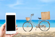 Summer sale concept, online shopping and devlivery, smartphone in girlhand with bicycle with jute bag over blurred beach background, summer outdoor day light