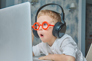 Cute child in red glasses and headphones has fun communicating online via video link using a laptop. The concept of modern means of communication and communication at a distance