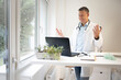 © epiximages - handsome doctor with white coat and blue shirt and stethoscope stands behind high table and works on laptop and has web conference or meeting