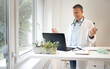 © epiximages - handsome doctor with white coat and blue shirt and stethoscope stands behind high table and works on laptop and has web conference or meeting