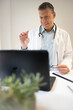 © epiximages - handsome doctor with white coat and blue shirt and stethoscope stands behind high table and works on laptop and has web conference or meeting