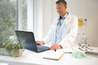 © epiximages - handsome doctor with white coat and blue shirt and stethoscope stands behind high table and works on laptop and has web conference or meeting
