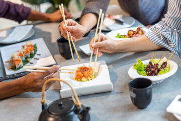 details of a table at the Japanese restaurant, hands of a multiethnic group with sticks picking up fusion food, seafood, sushi and sashimi