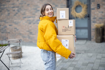 Cheerful woman in bright jacket with parcels in front of her house, coming home with goods purchased online. Shopping online and delivering goods concept