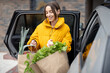 Woman in yellow jacket taking paper bags full of fresh groceries from a car, arriving home with a purchases. Shopping healthy food in eco-packaging