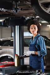 young african american mechanic standing underneath car, holding motor oil bottle and looking at camera in garage