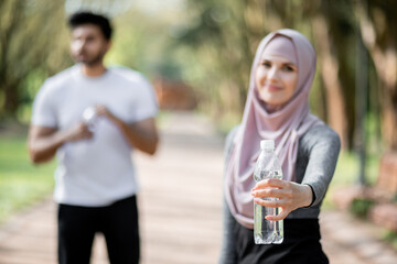 Wall Mural - Blur background of young woman in hijab and muslim man standing at green city park with fresh water. Focus on female hand with bottle of water. Refreshing concept.