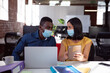© wavebreak3 - Diverse male and female colleague in face masks sitting at desk discussing, using laptop and tablet