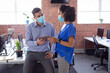 © wavebreak3 - Diverse male and female colleague in face masks looking at tablet and discussing standing in office