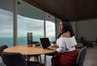 © Joseph De Frías - Latin woman sitting at the table in her apartment working from home with her laptop and smartphone in the background a large window with view to the sea. Remote working concept