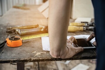 Carpenter working with wood furniture product preparation using  hand tools and machine table station while using mobile phone