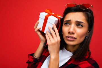 Closeup photo of beautiful emotional young brunet woman isolated on red background wall wearing white casual t-shirt and stylish red and black shirt holding white gift box with red ribbon